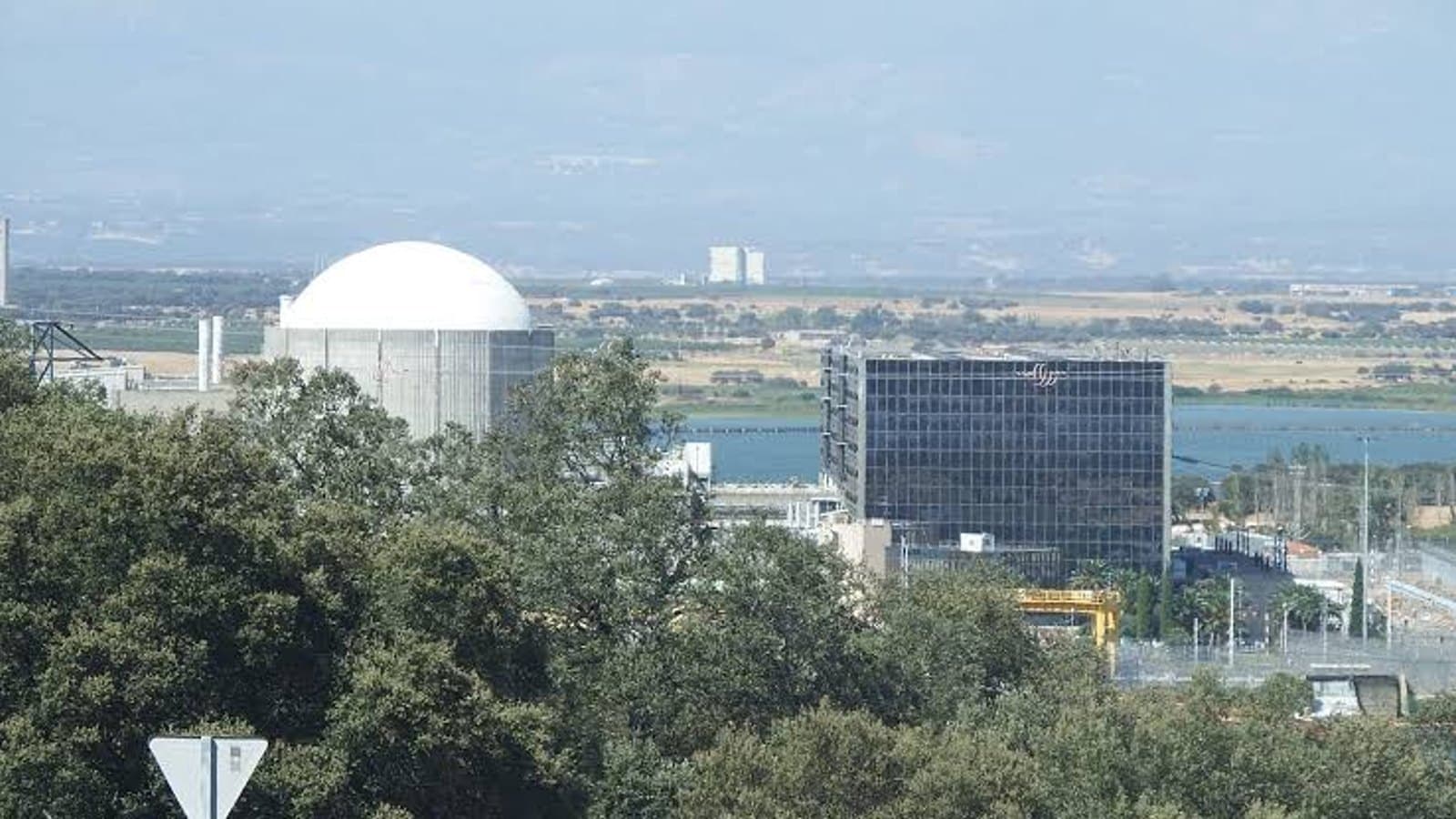 The domed reactor building of the Almaraz nuclear plant seen across trees and a reservoir, next to a glass fronted building.