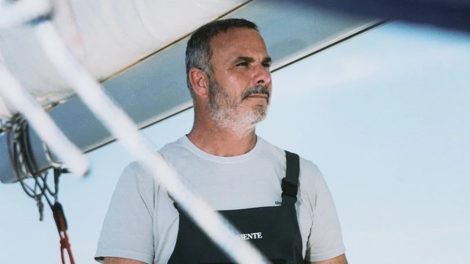 Ángel León de pie en un velero bajo la botavara, con camiseta blanca y peto oscuro de Aponiente, mirando hacia un lado.