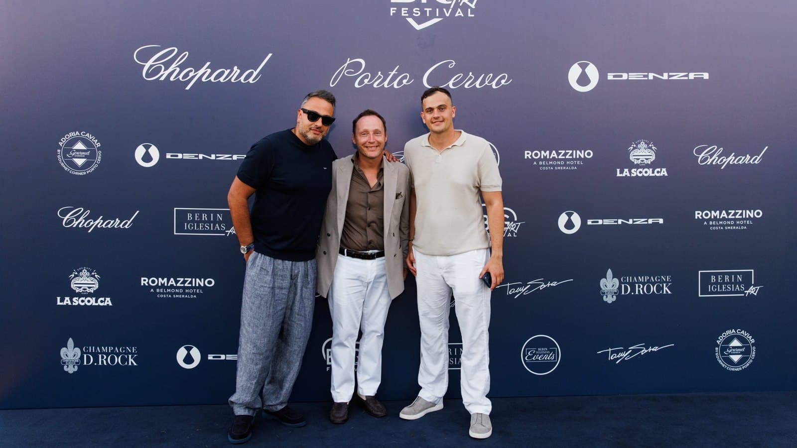 Tres hombres posan juntos ante un fondo de photocall de BIG ART Festival con los logotipos de Chopard, Porto Cervo y Denza.