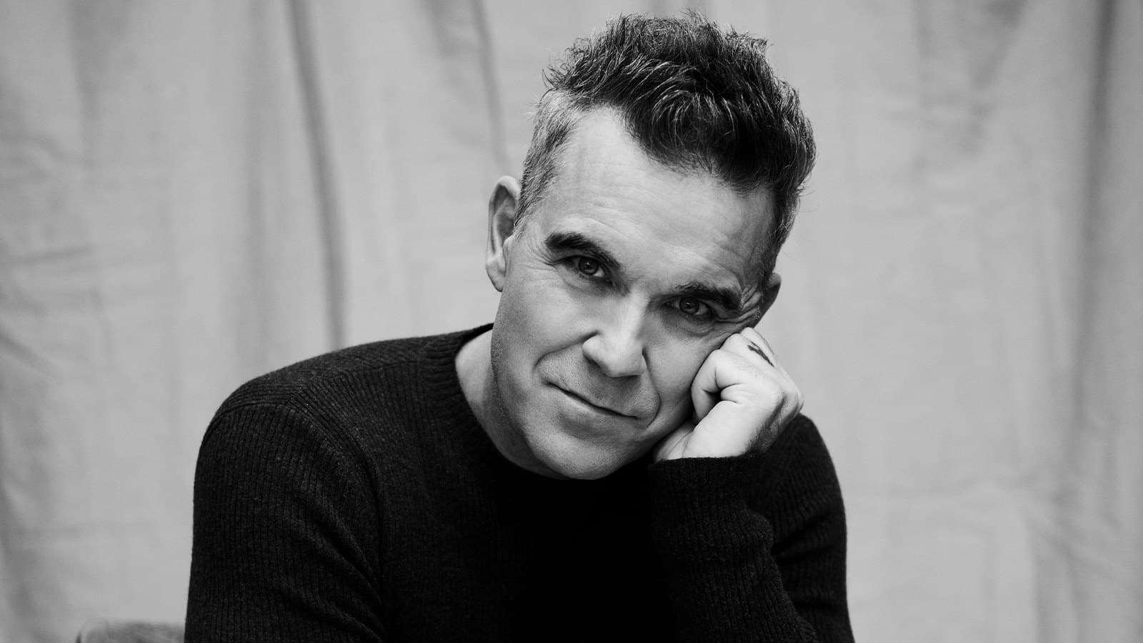 Robbie Williams photographed in a dark knit sweater, resting his cheek on his hand, in black and white.