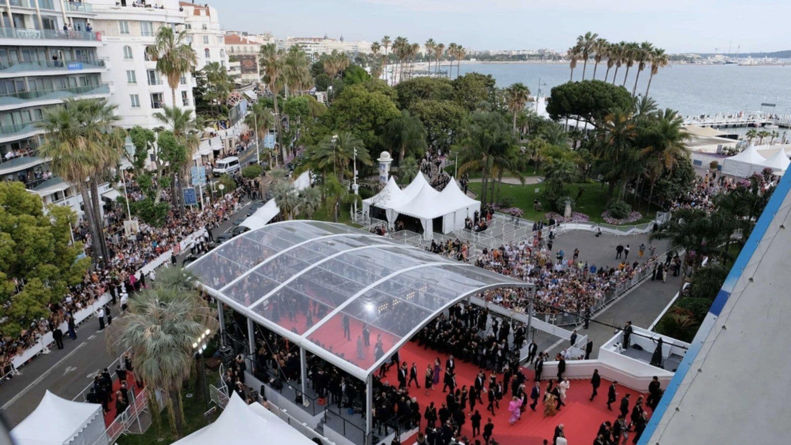 An aerial view of the Cannes red carpet under its glass canopy, with guests arriving, crowds behind barriers along the street and the sea beyond.