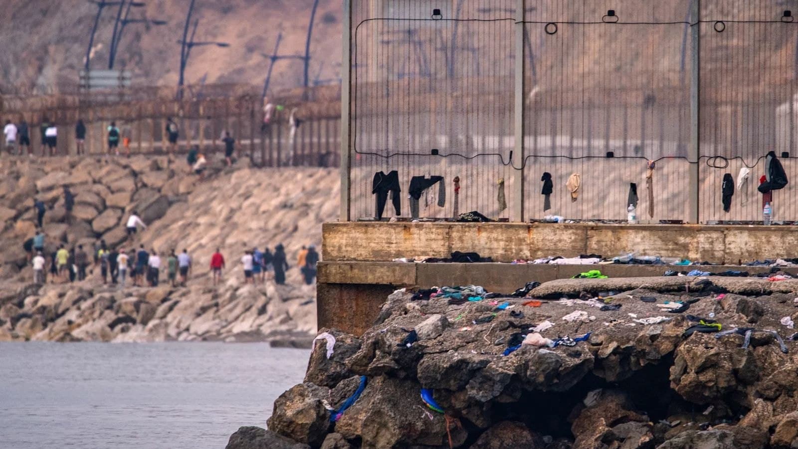 Ropa abandonada cuelga de una valla fronteriza sobre un dique de rocas en Ceuta, con personas caminando entre las rocas al fondo.