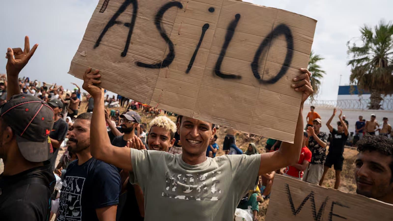 A young man smiles while holding up a cardboard sign reading Asilo above a large crowd seated outdoors.