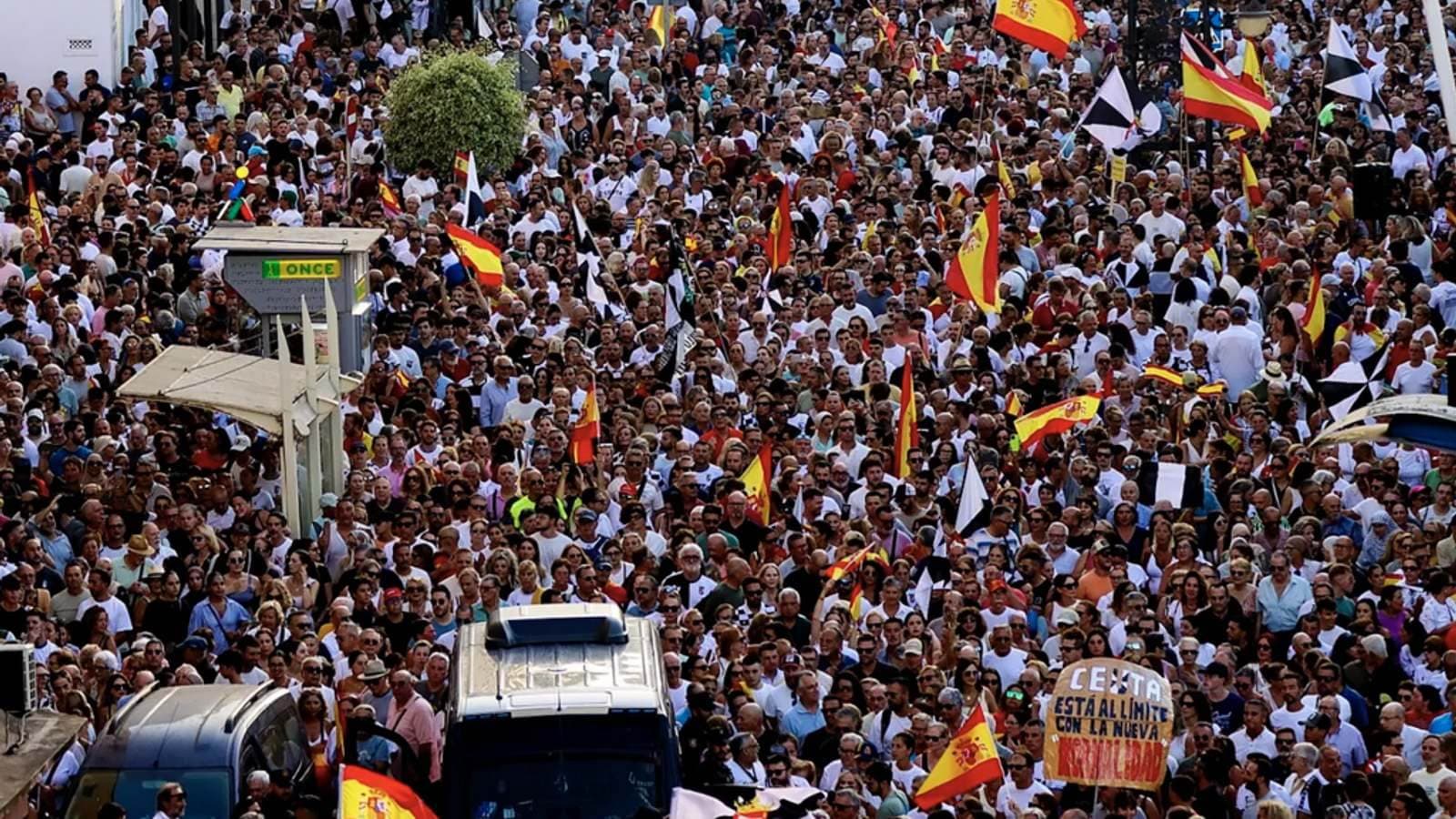 Una multitud densa llena una calle ondeando banderas españolas, con un cartel escrito a mano que dice Ceuta está al límite con la nueva normalidad.