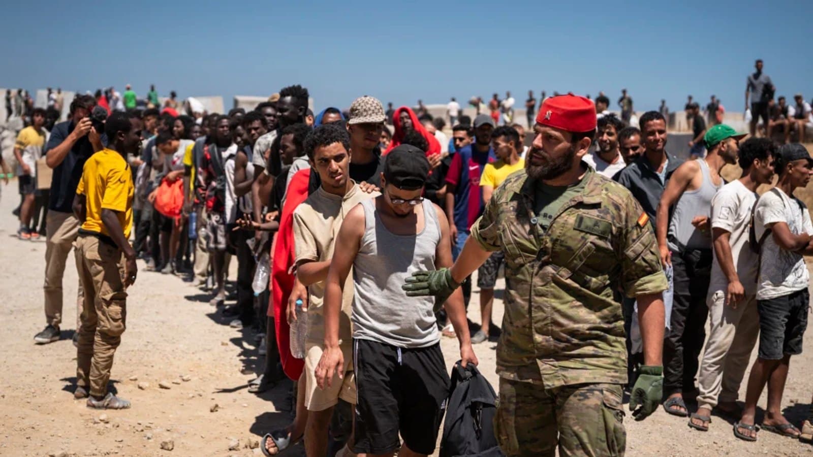 Un soldado de la Legión española con boina roja guía una larga fila de jóvenes que esperan al aire libre bajo un cielo despejado.