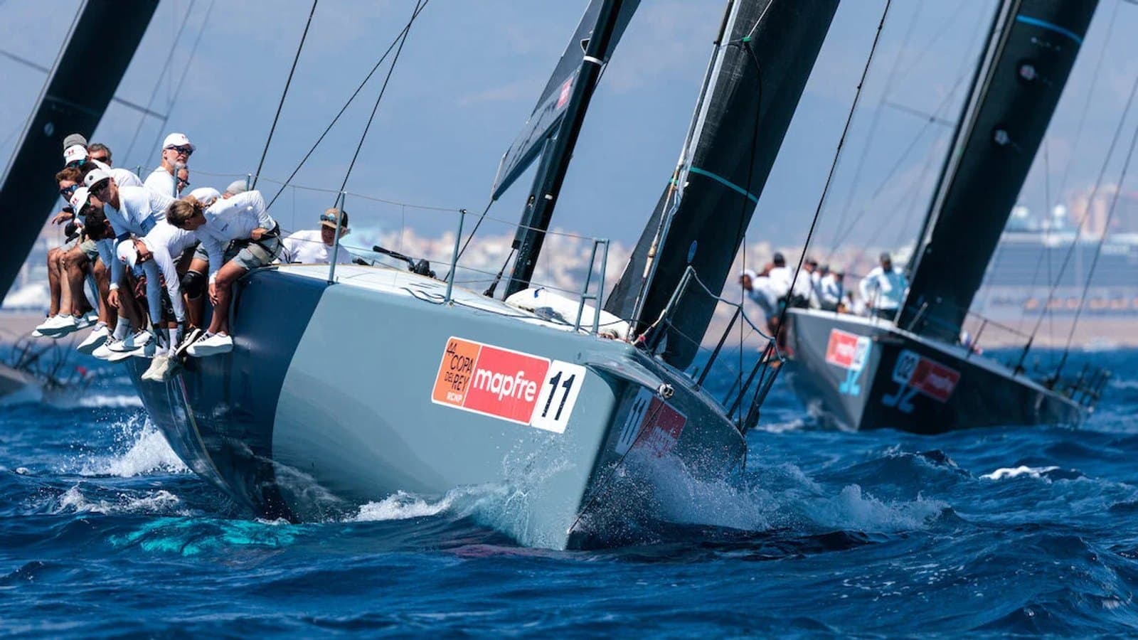 La tripulación de un TP52 se inclina sobre la borda mientras el barco escora con fuerza al cortar una ola, con un rival muy cerca por detrás.