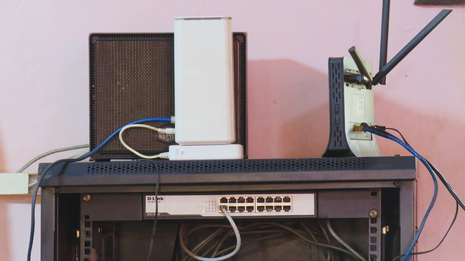 A front facing shot of a black wall mounted network rack containing a 16 port network switch with several connected Ethernet cables. Placed on top of the rack are a white wireless router, a black modem with dual antennas. The equipment is set against a light pink wall, with a tangle of black and grey cables visible inside. Fotografia de archivo.