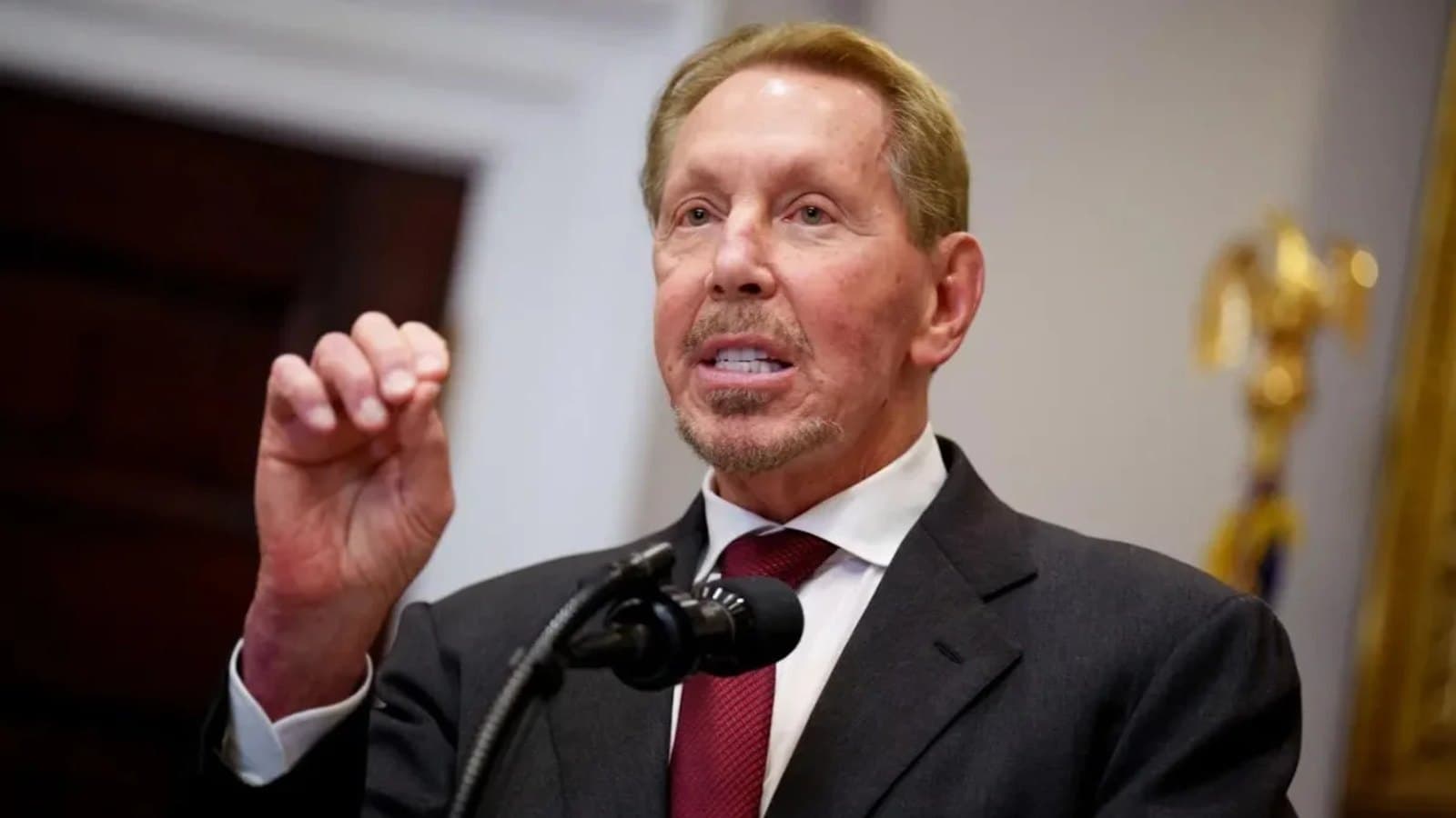 Larry Ellison gestures with a raised fist while speaking at a lectern with a microphone, in a dark suit and burgundy tie.