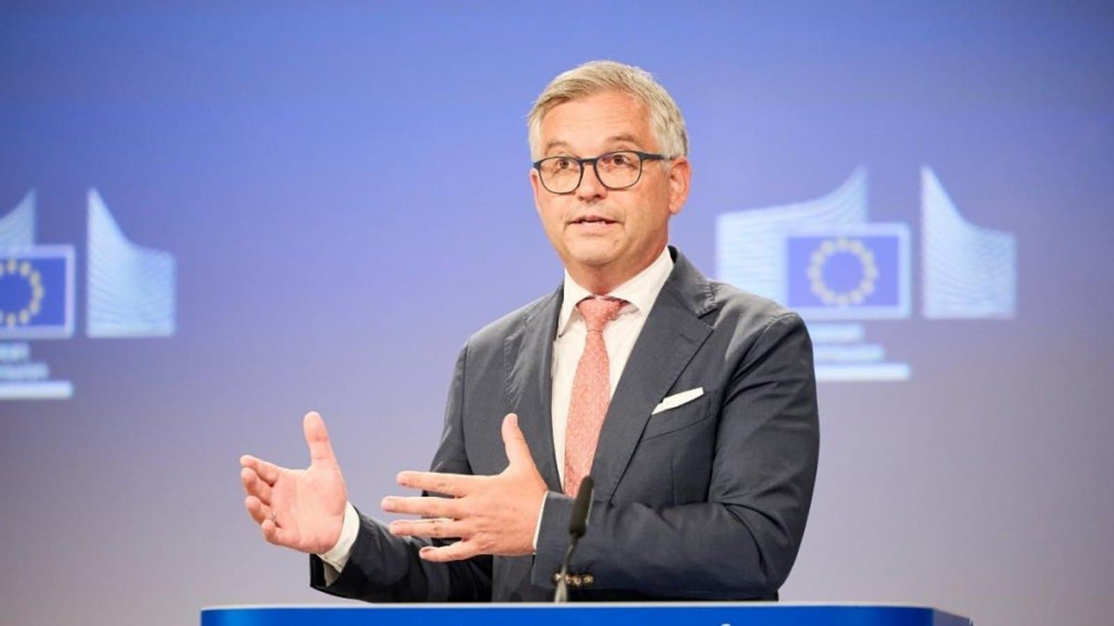 A man in a dark suit and pink tie gestures with both hands while speaking at a lectern marked European Commission, with EU emblems on the wall behind him.