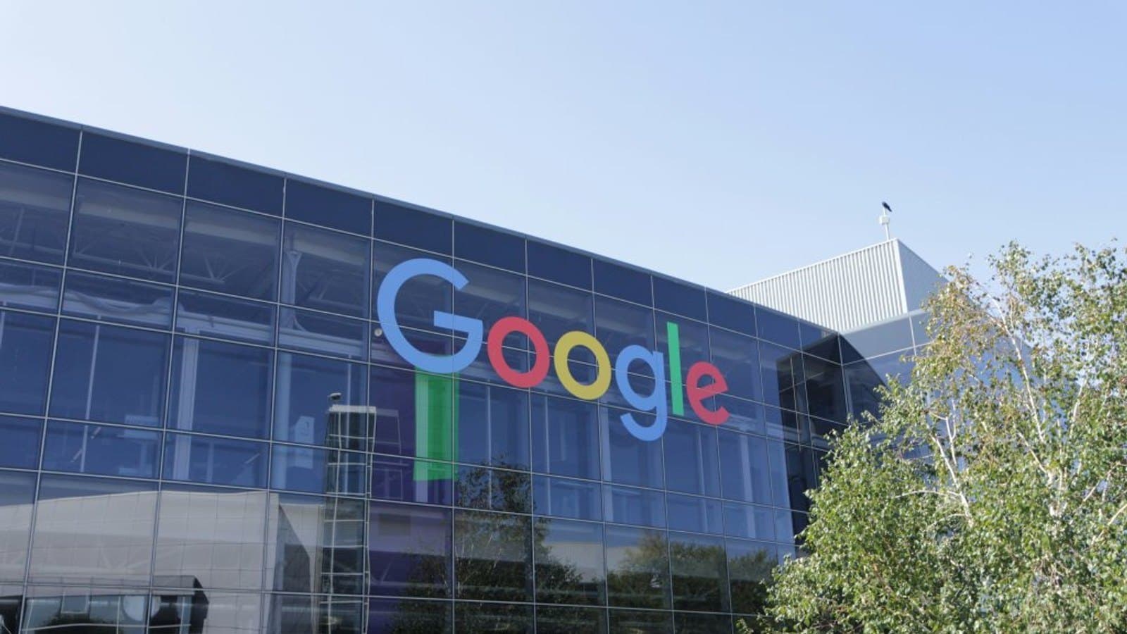 El logotipo de Google en la fachada acristalada de un edificio de oficinas bajo un cielo despejado, con árboles delante.
