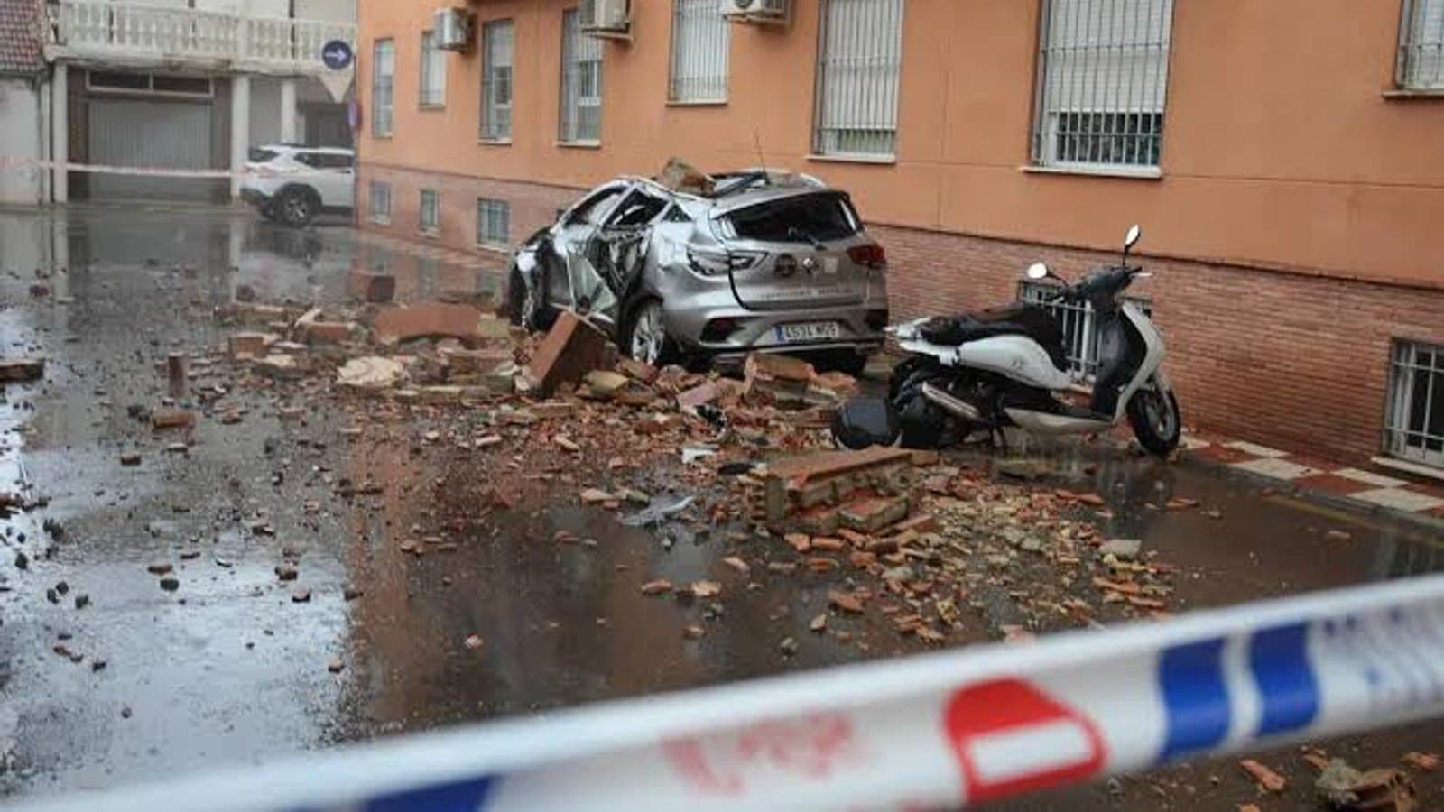 Un coche destrozado bajo ladrillos caídos junto a una moto, tras cinta policial, con un edificio de viviendas dañado encima.