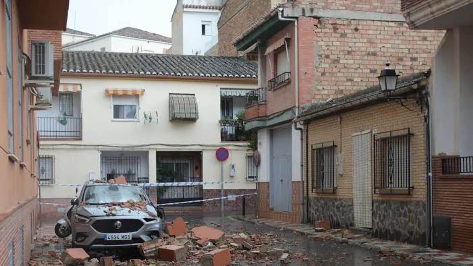 Un coche cubierto de tejas rotas y escombros, acordonado con cinta policial en una calle de Alhendín, con la fachada de un edificio dañado al fondo.