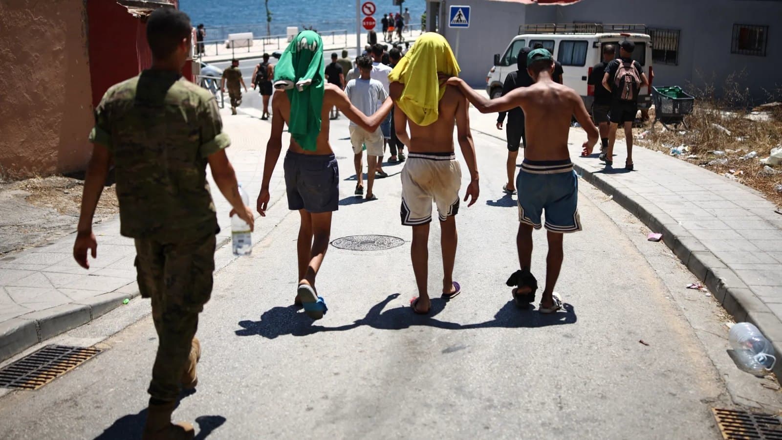 Un soldado camina detrás de tres jóvenes con camisetas sobre la cabeza para protegerse del sol, en una calle sobre el paseo marítimo de Ceuta.