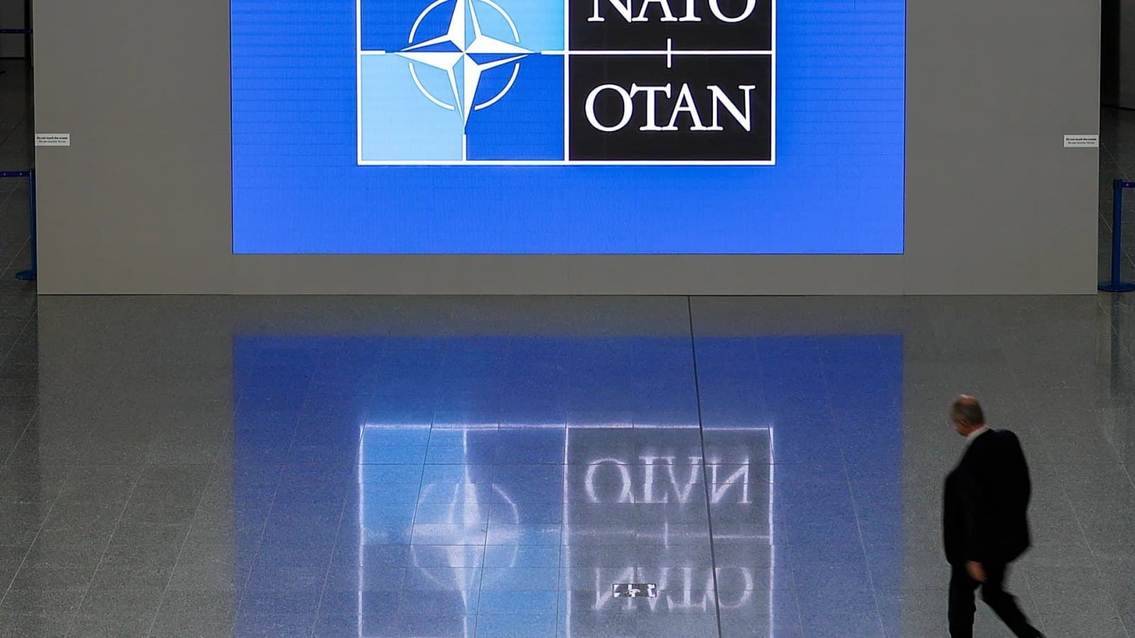 A man in a dark suit walks past an illuminated NATO OTAN logo display in a marble hallway, reflected in the polished floor.