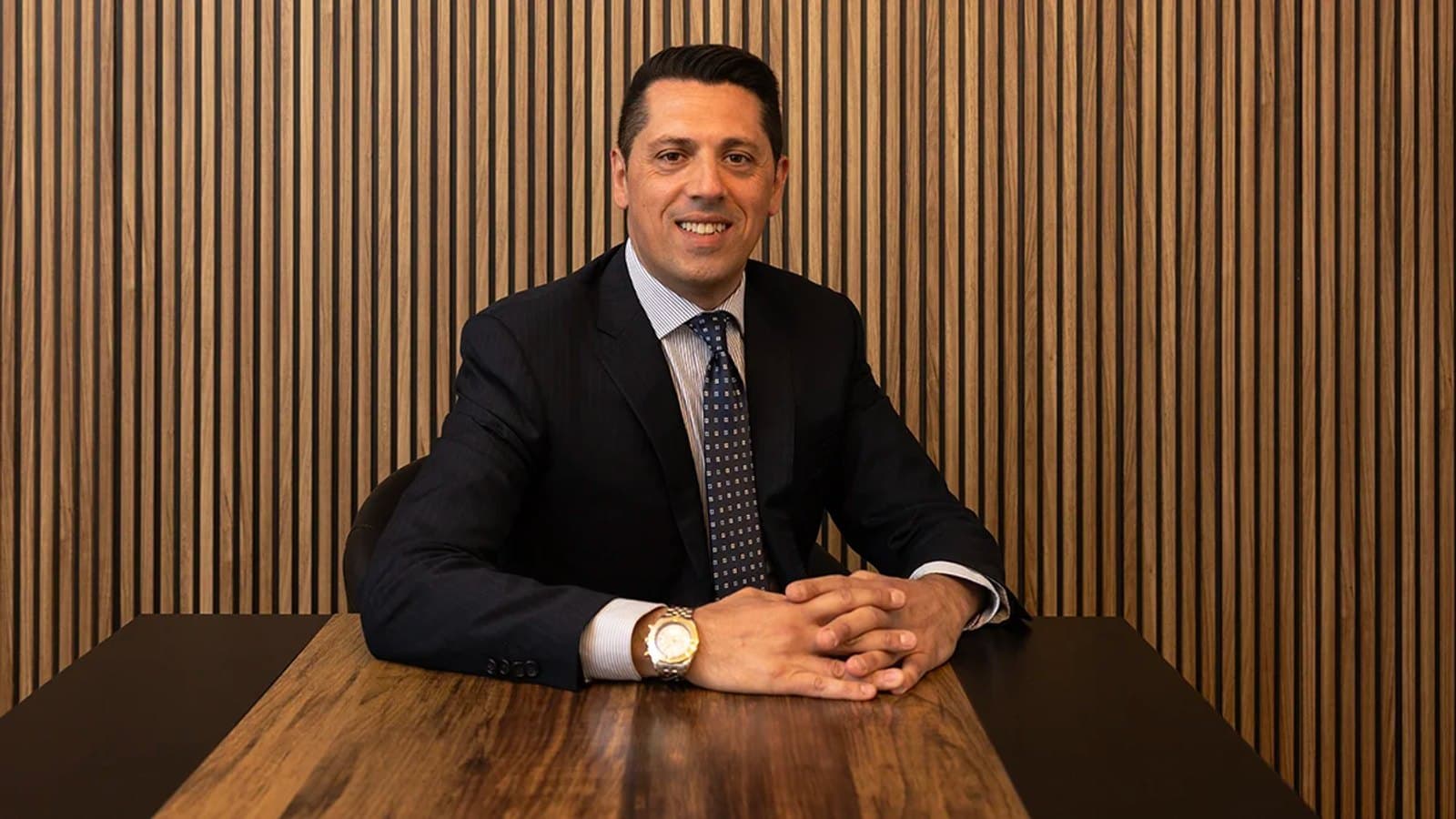 Mauro Povia, con traje y corbata oscuros, sentado a una mesa de madera con las manos entrelazadas ante una pared de paneles de madera acanalados.