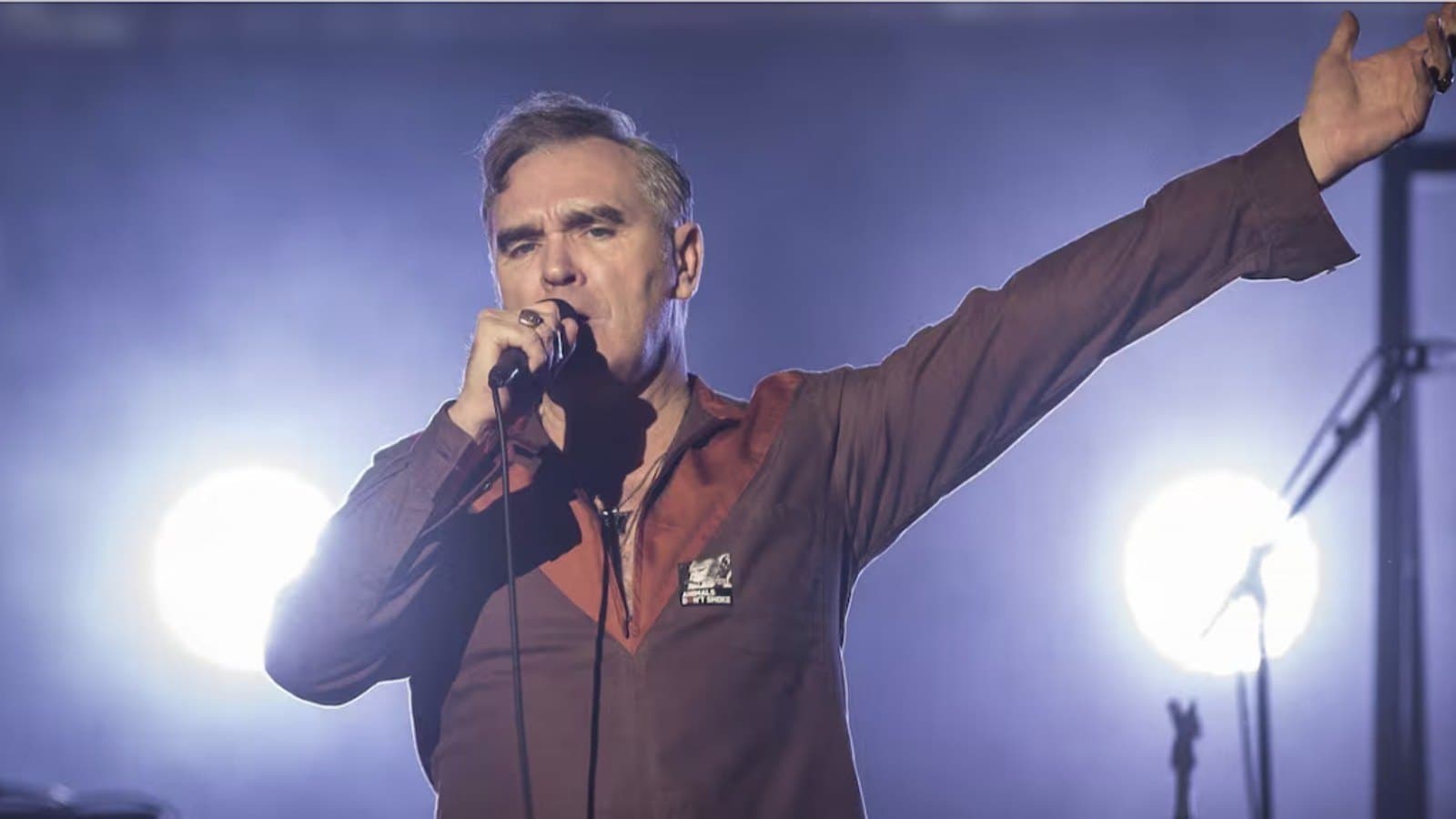 A singer in a dark red shirt holds a microphone to his mouth and throws one arm out to the side, lit from behind by two white stage lights.