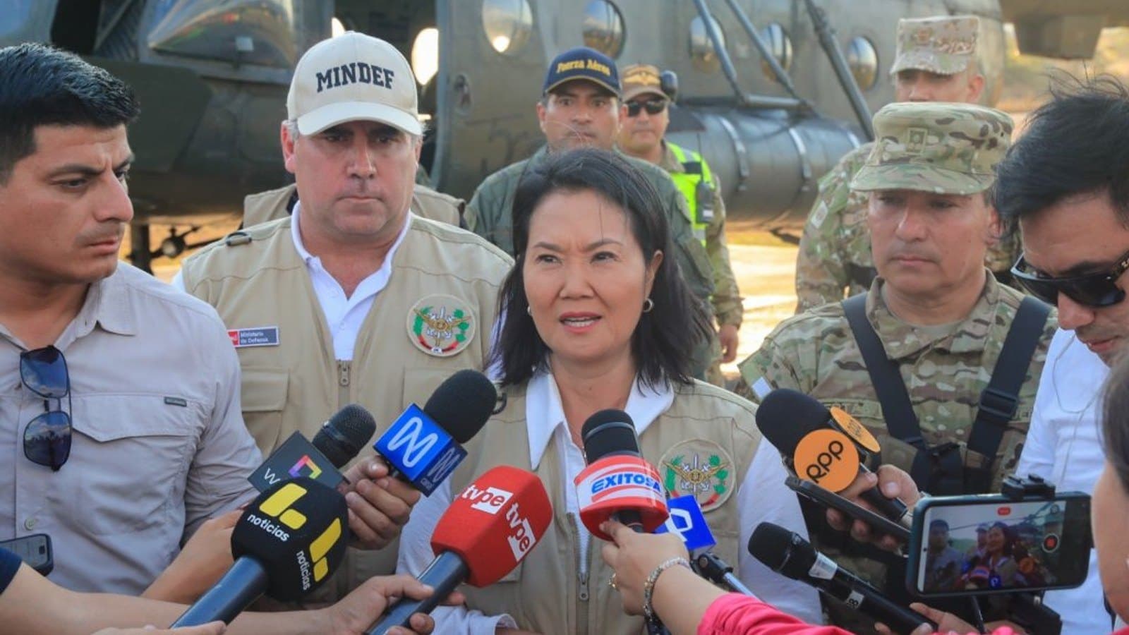 Una mujer con chaleco de campaña habla ante un grupo de micrófonos de televisión, con personal uniformado y un helicóptero militar detrás.