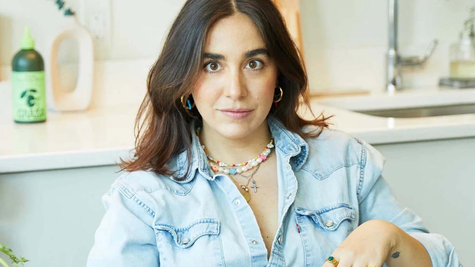 Paula Casado con camisa vaquera, sentada en una cocina con collares superpuestos y aros dorados.