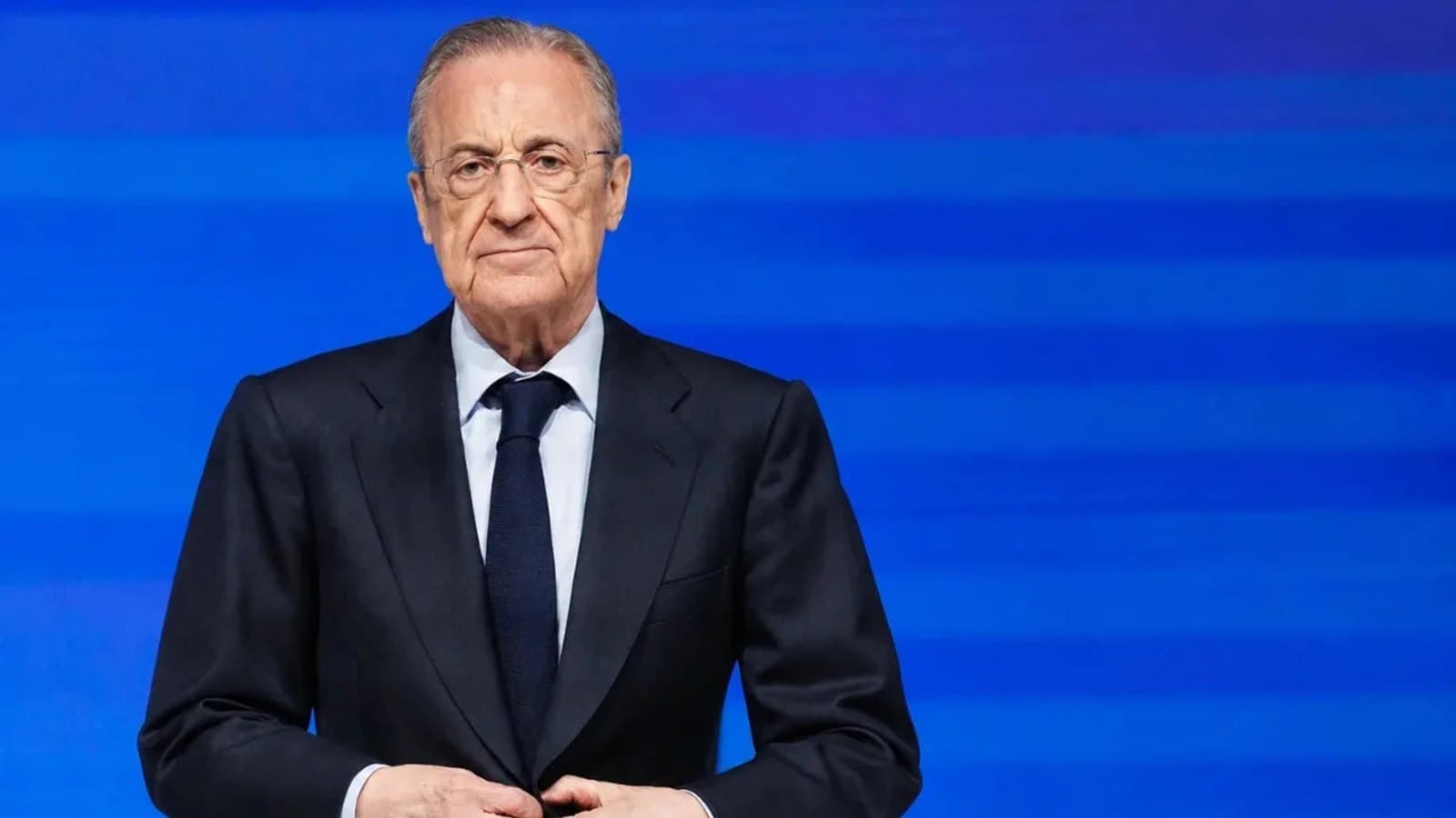 Florentino Pérez con traje oscuro y gafas, de pie con las manos entrelazadas ante un fondo azul de rayas.