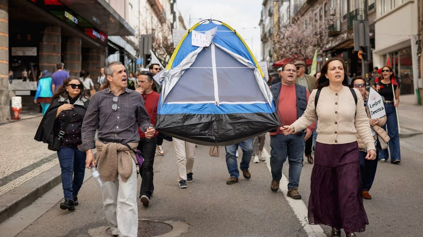 Protesters march down a city street carrying a camping tent overhead, with a sign reading Lembrar Democracia among the crowd.