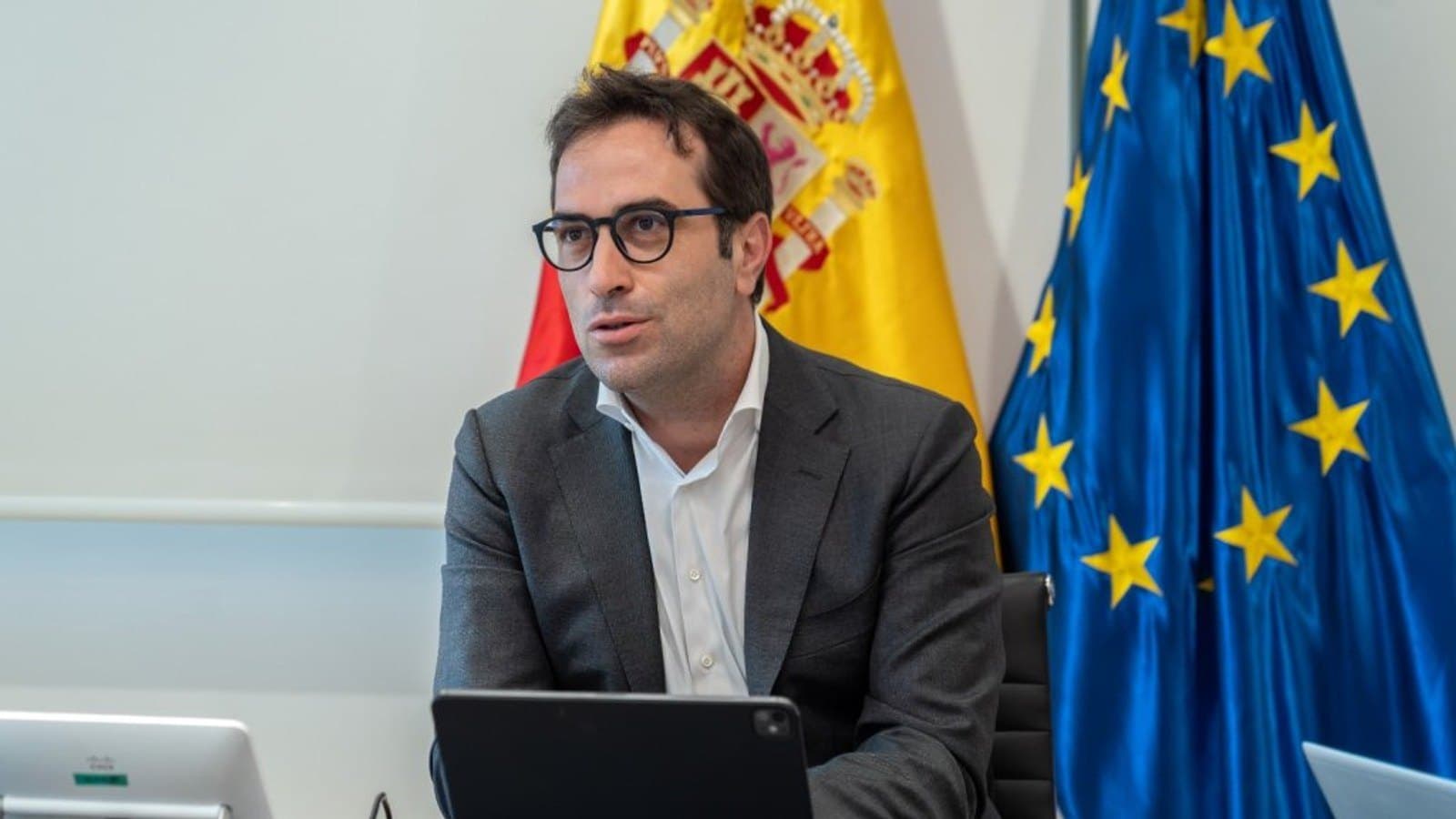 Un hombre con gafas y traje gris habla ante una mesa con una tableta, sentado delante de las banderas de España y de la Unión Europea.