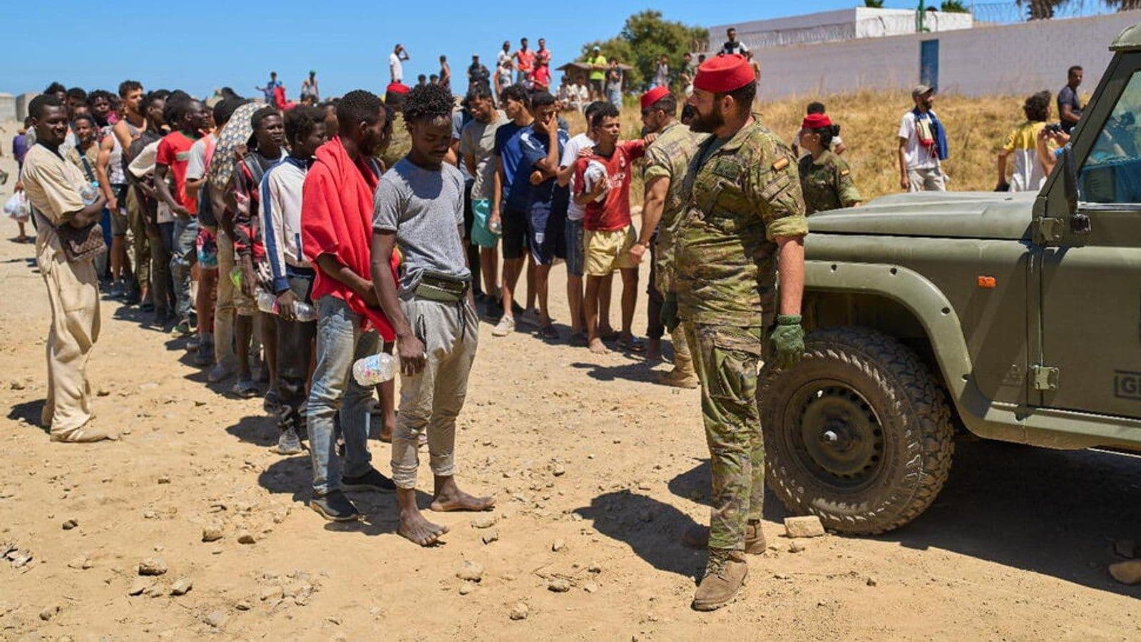 A line of young men, most carrying little more than water bottles, waits on dry ground beside a Spanish soldier in camouflage and a military vehicle, with more people gathered on a slope behind them.