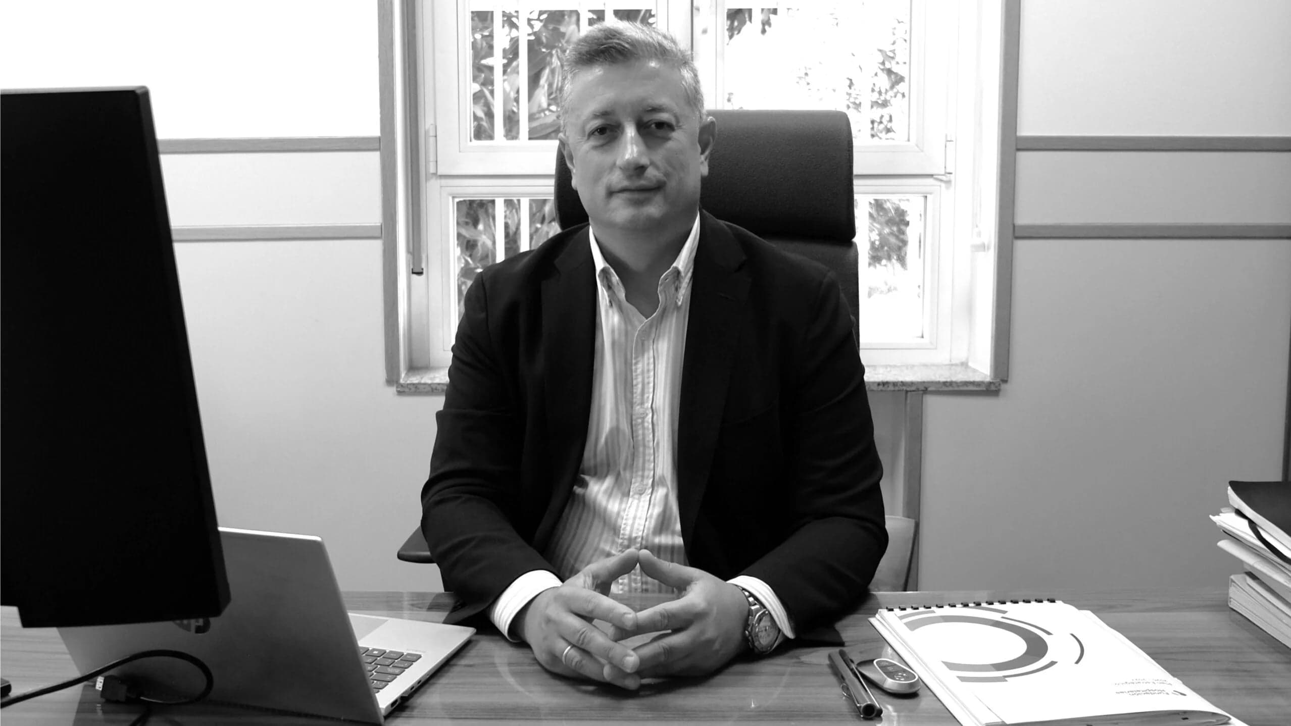 Retrato en blanco y negro de un hombre con traje sentado en un despacho junto a un ordenador y documentos