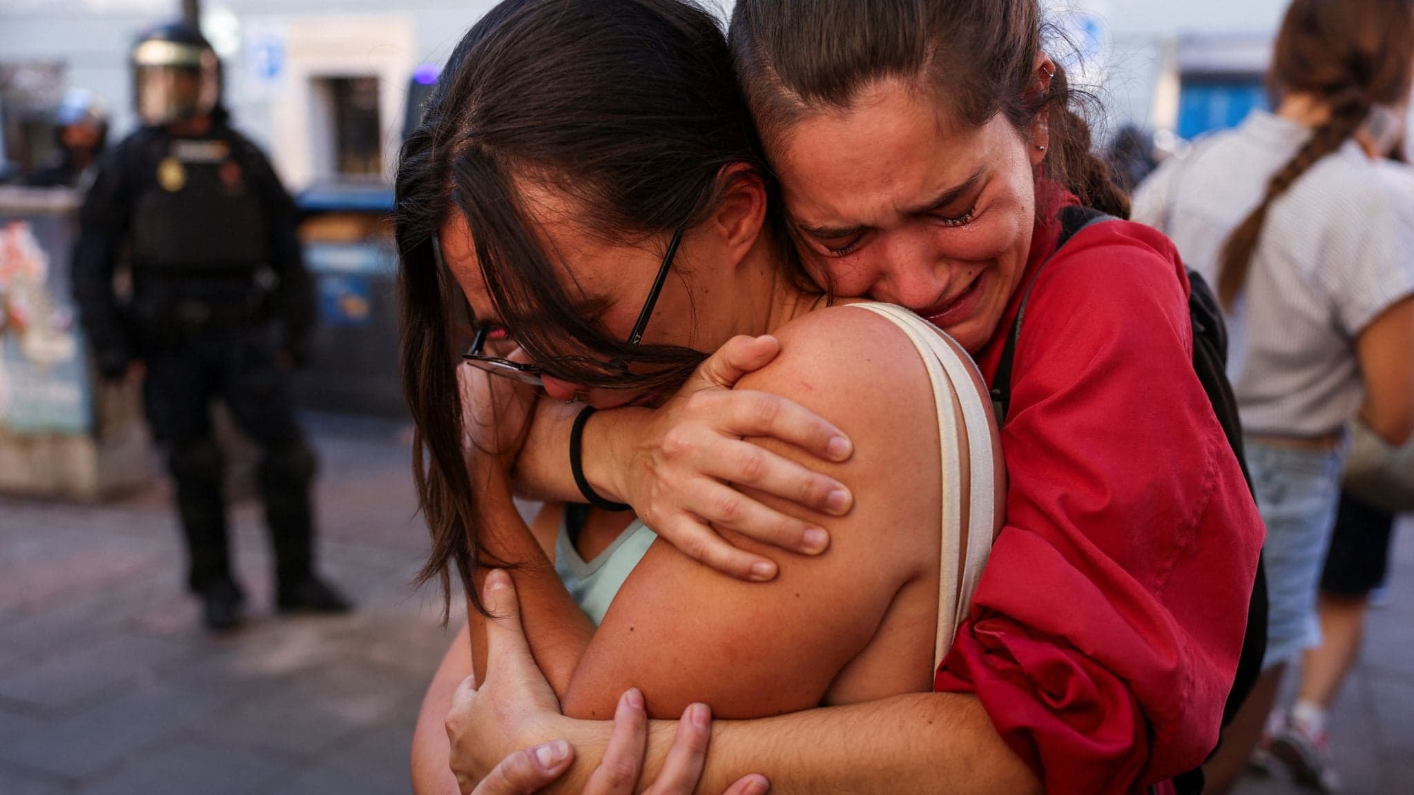 Two women embrace in tears during a protest in Madrid as a riot police officer stands watch in the background