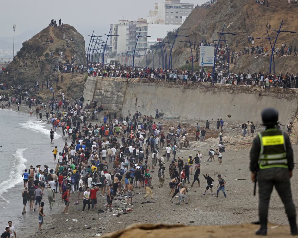 Una multitud se agolpa junto al muro fronterizo y la playa de Ceuta mientras un agente de seguridad observa la escena