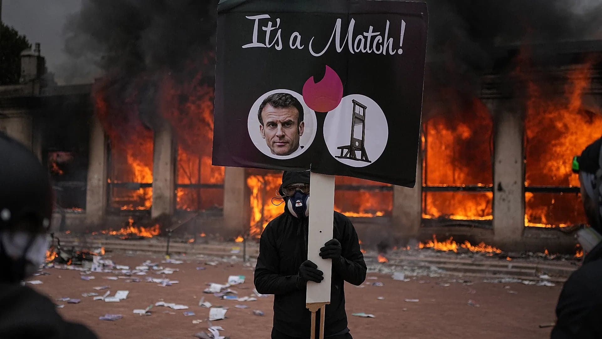 A protester holds a sign mocking French President Emmanuel Macron in front of a burning building during cost of living protests in France