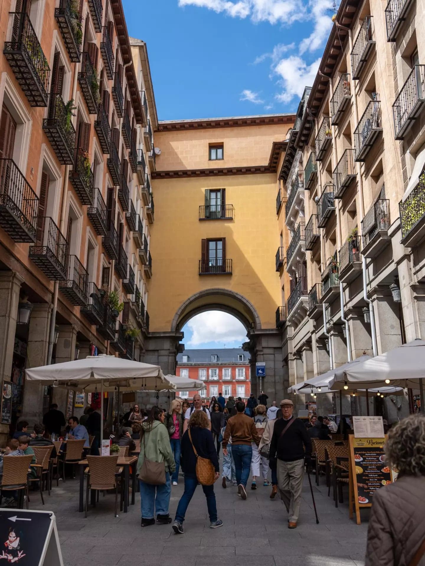 Una calle peatonal de Madrid con terrazas y un arco entre edificios historicos, llena de gente paseando