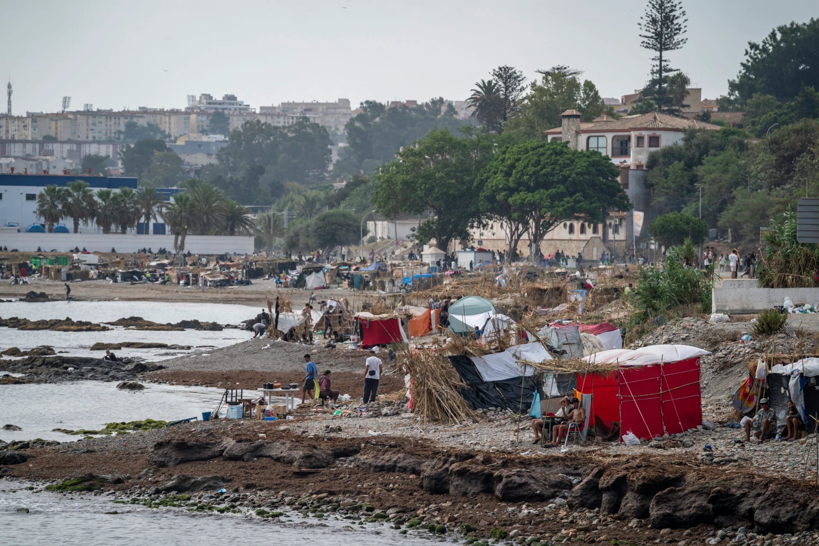 A makeshift migrant encampment with tents along the Ceuta coastline, with city buildings in the background