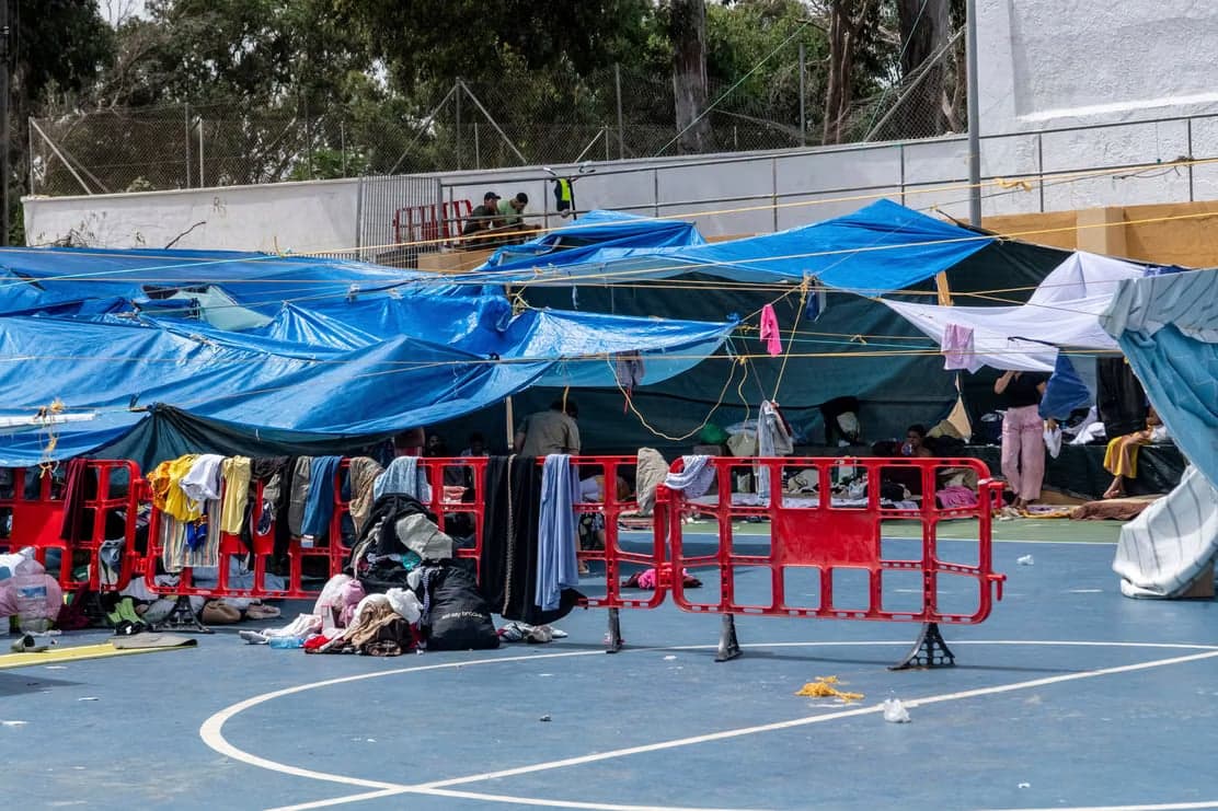 A makeshift migrant camp of tarps and tents set up on a sports court in Ceuta, with clothing hung to dry along red barriers