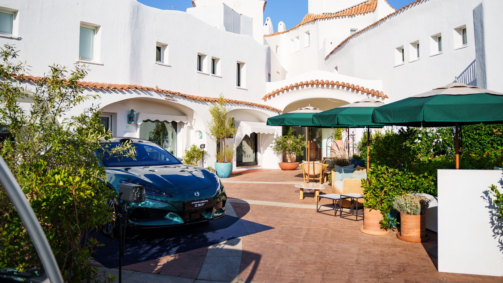 Un Denza Z9 GT verde oscuro aparcado en el patio encalado de Romazzino, A Belmond Hotel, bajo un cielo azul despejado.