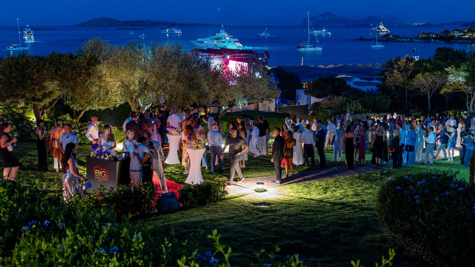 Invitados se reúnen en los jardines iluminados de Romazzino por la noche, con yates anclados en la bahía al fondo.