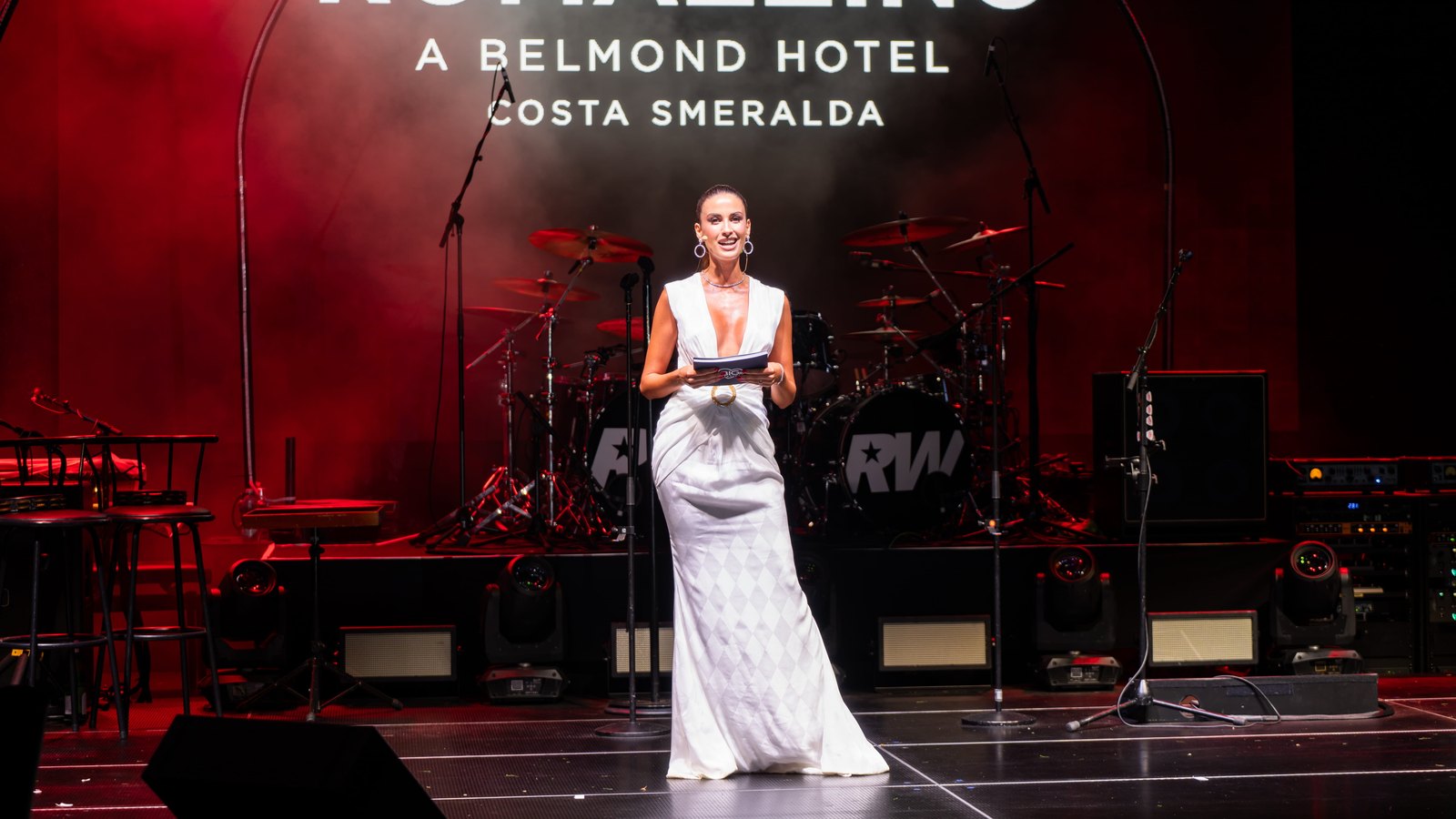 Eleonora Incardona con un vestido blanco sobre el escenario, ante un fondo de Romazzino, A Belmond Hotel iluminado en rojo.