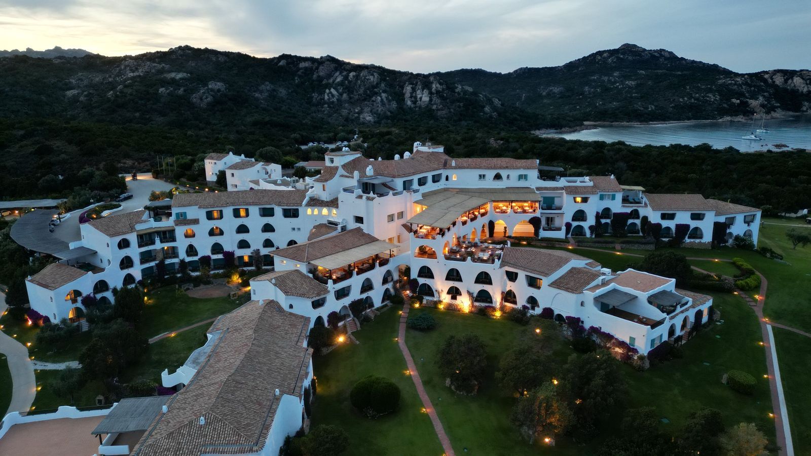 Vista aérea al atardecer del hotel Romazzino, un complejo blanco de poca altura con tejados de teja, senderos iluminados y la costa de la Costa Esmeralda al fondo.